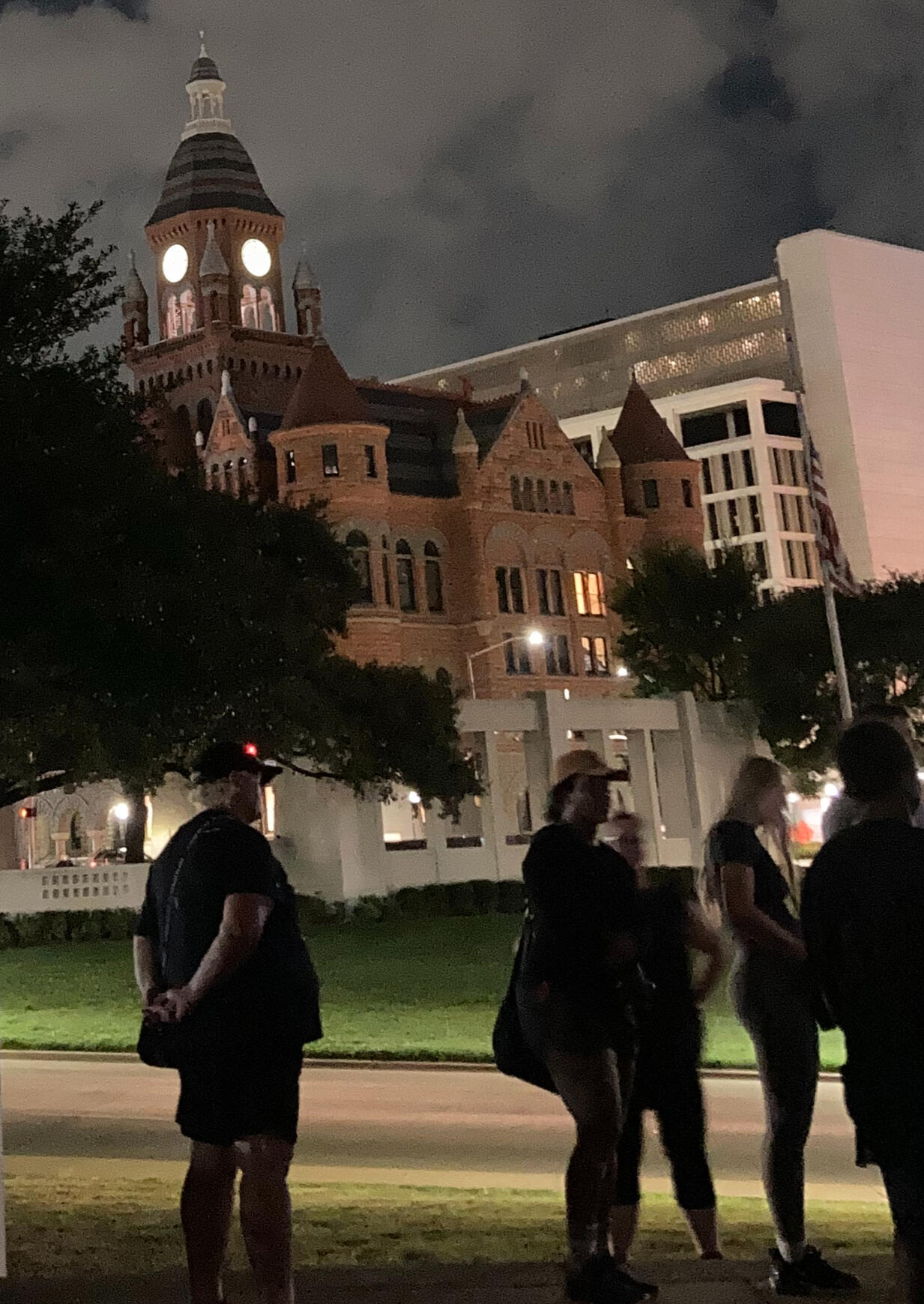 A photo from one of our true crime walking tours showing the big red court house at dusk the clock tower is lit in a warm glow a crowd of people gather to listen to our guide tell his story