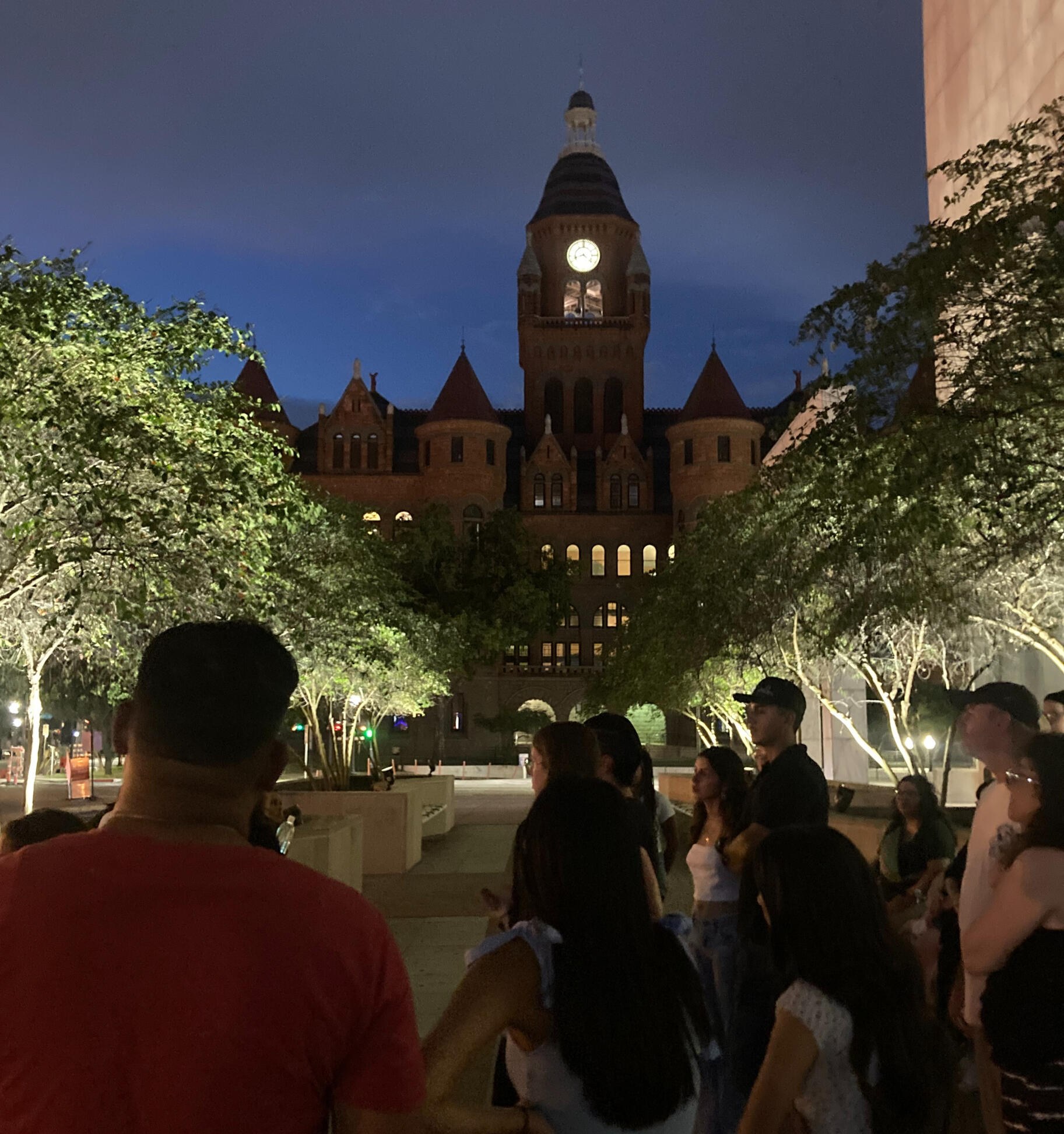 A photo from one of our true crime walking tours showing the big red court house at dusk the clock tower is lit in a warm glow a crowd of people gather to listen to our guide tell his story