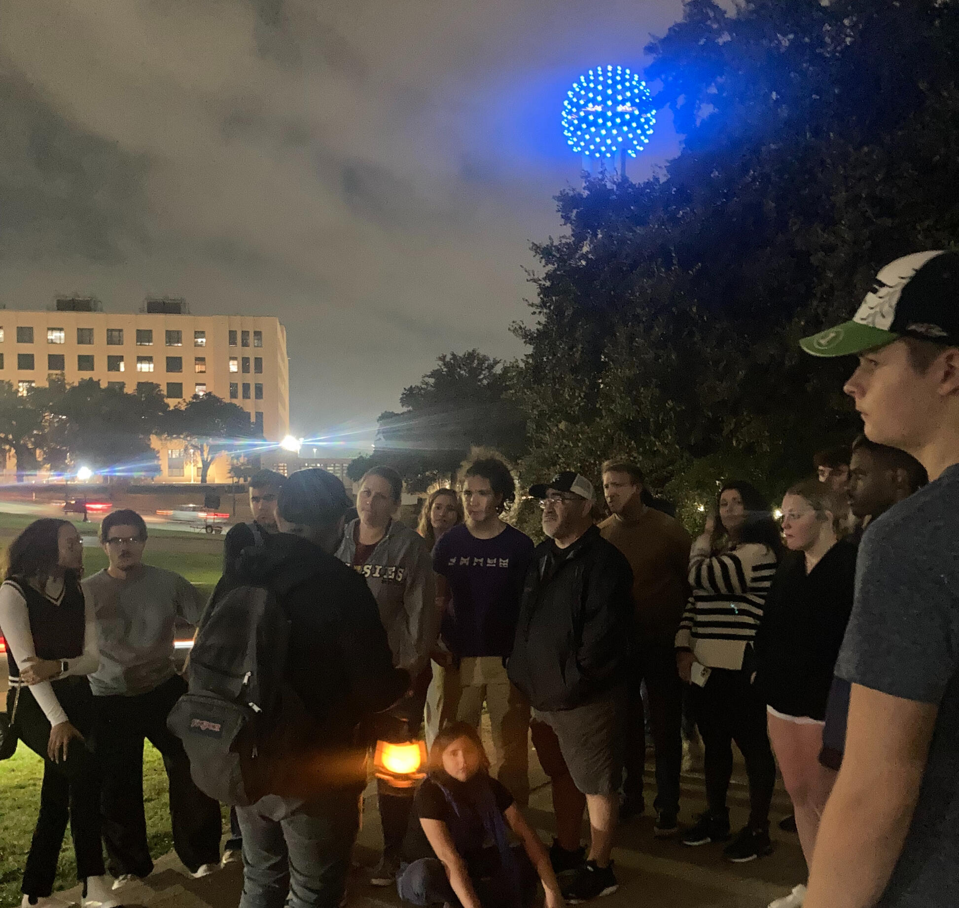 A photo from one of our true crime walking tours showing reunion tower at dusk lit in blue neon a crowd of people gather to listen to our guide tell his story