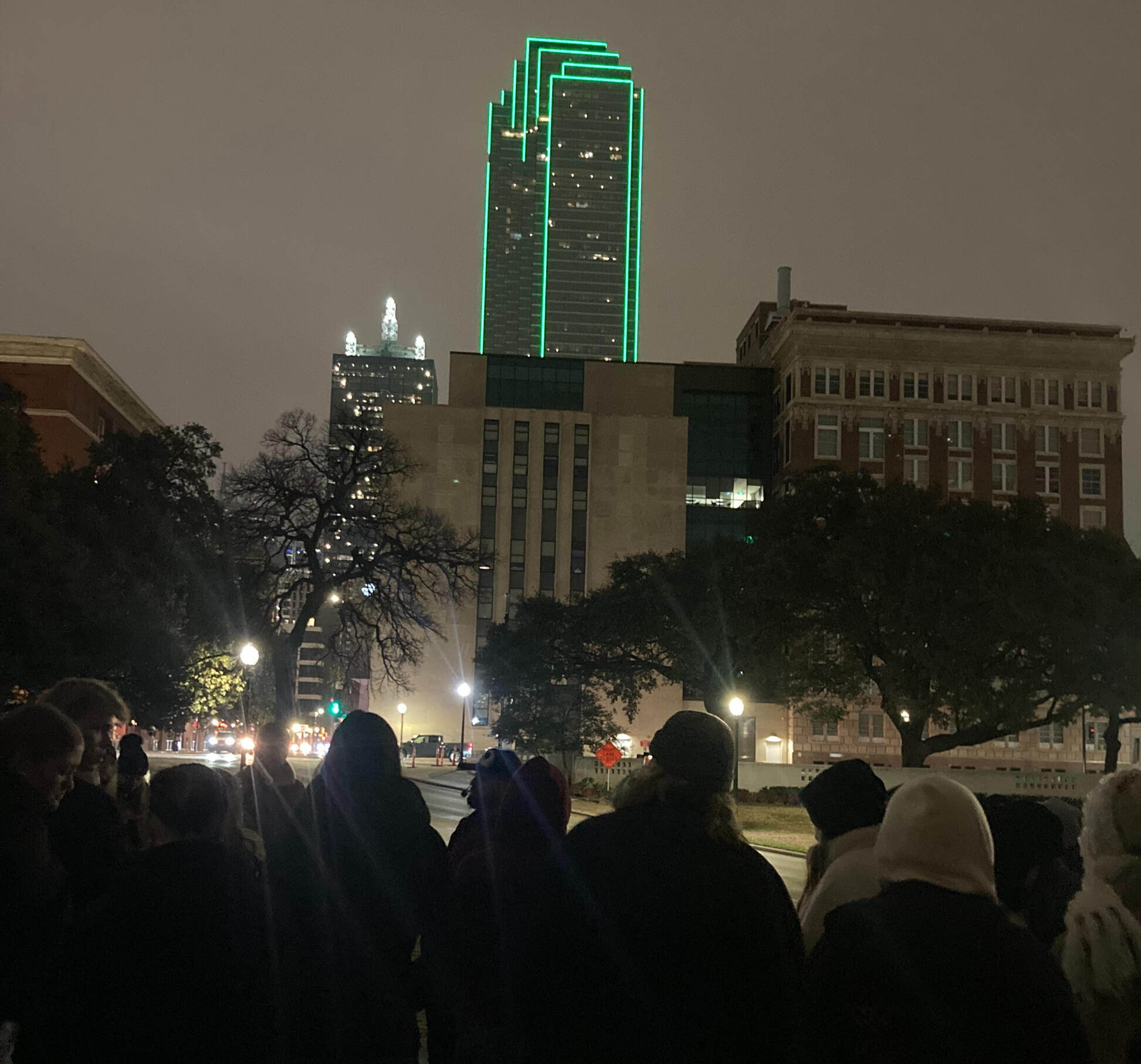 A photo from one of our true crime walking tours showing the bank of america tower at dusk lit in green neon a crowd of people gather to listen to our guide tell his story